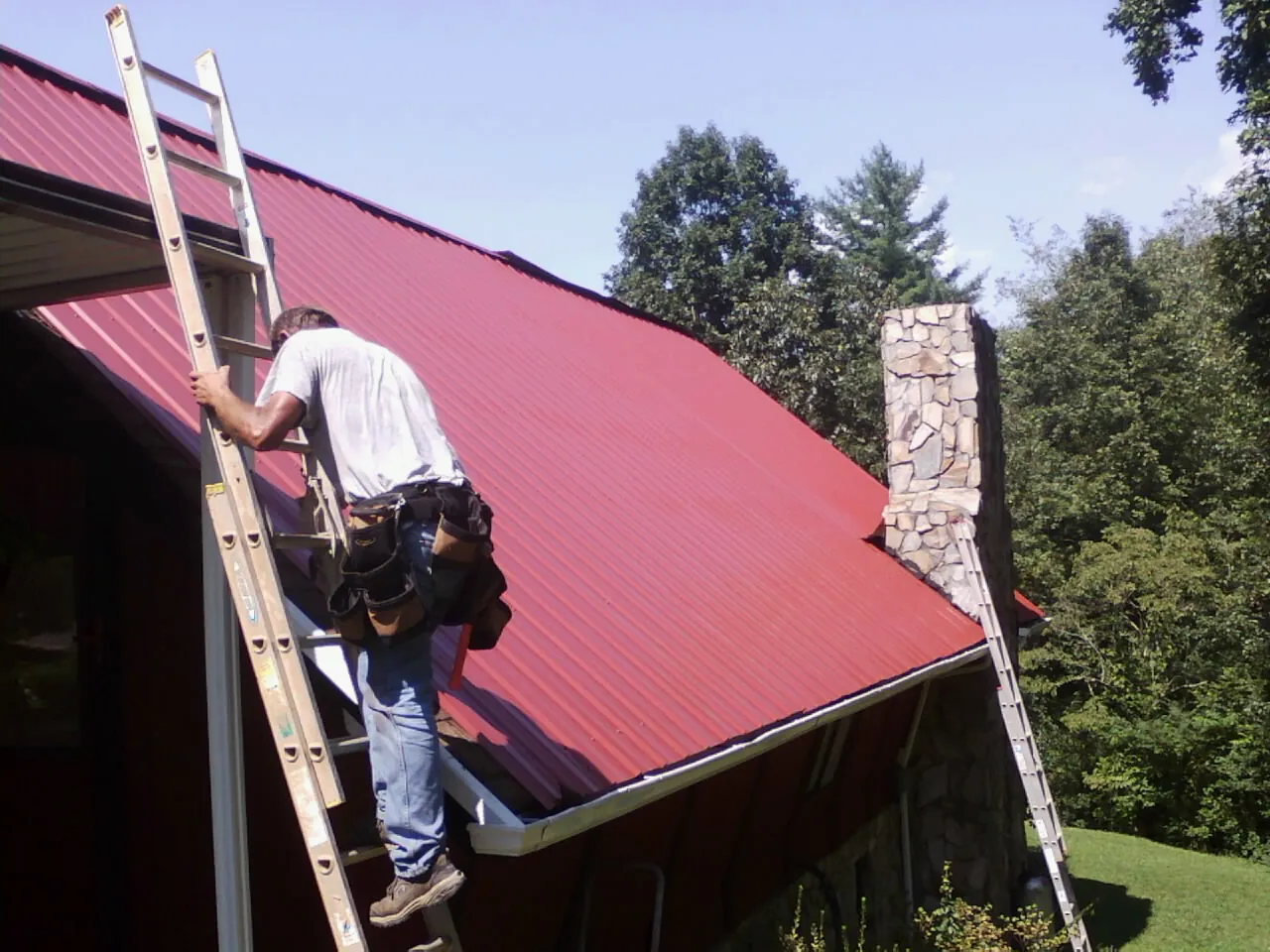 Roofer on a ladder beside a red metal roof and stone chimney — Soffit & Fascia Repair by Roofing Brothers in Gallion